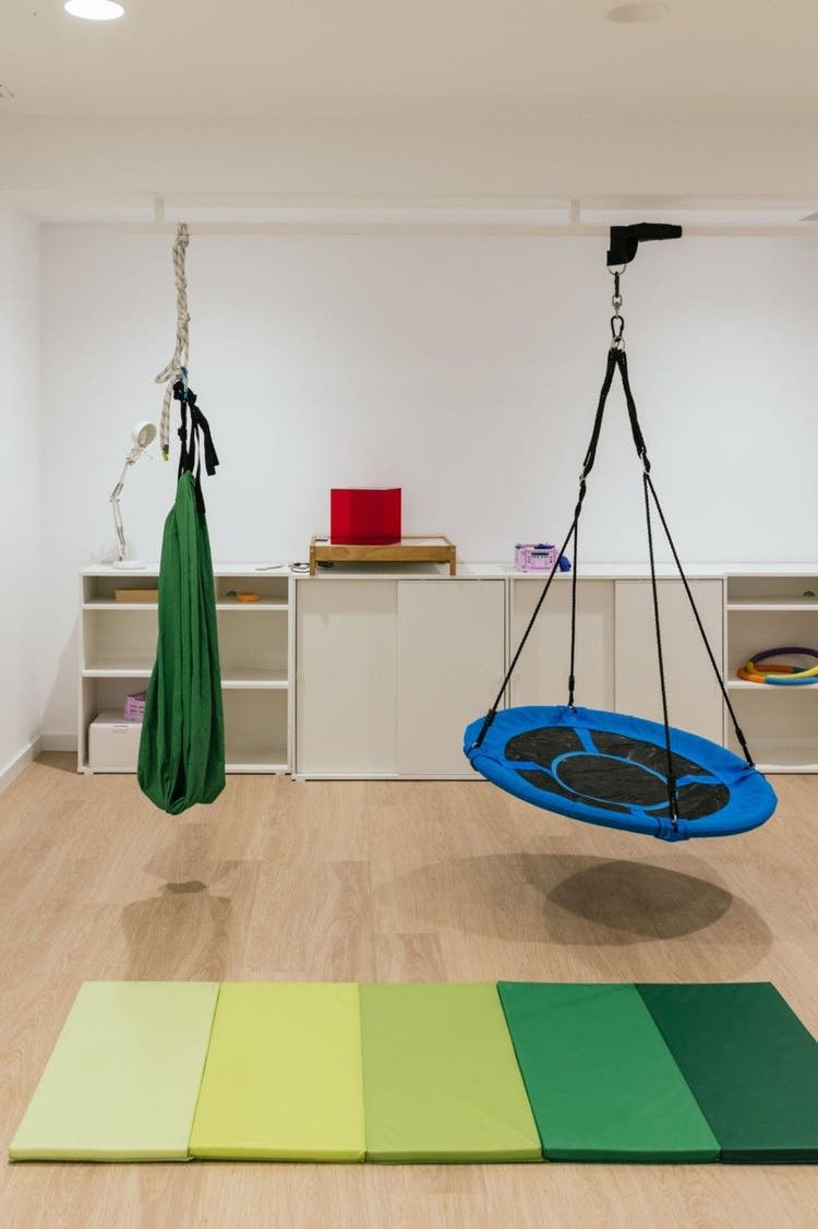 An empty sensory therapy room featuring a green pod swing and a blue platform swing suspended above a colourful floor mat.