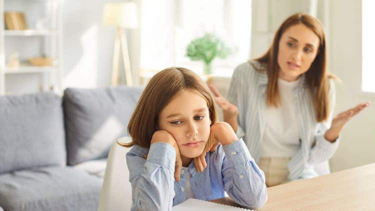 A sad young girl looking away while a frustrated woman gestures in the background.