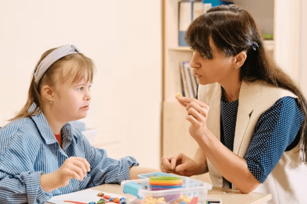 A female therapist speaking to a young girl while holding up a small token during a learning session.